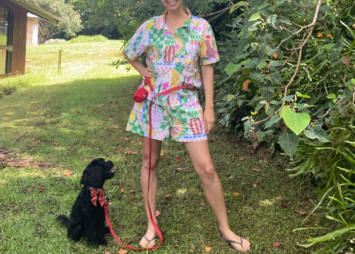 A woman stands on a grassy lawn wearing a colorful tropical print short-sleeve top and matching shorts, with a red leash and a small black dog beside her. Trees and garden foliage fill the background.